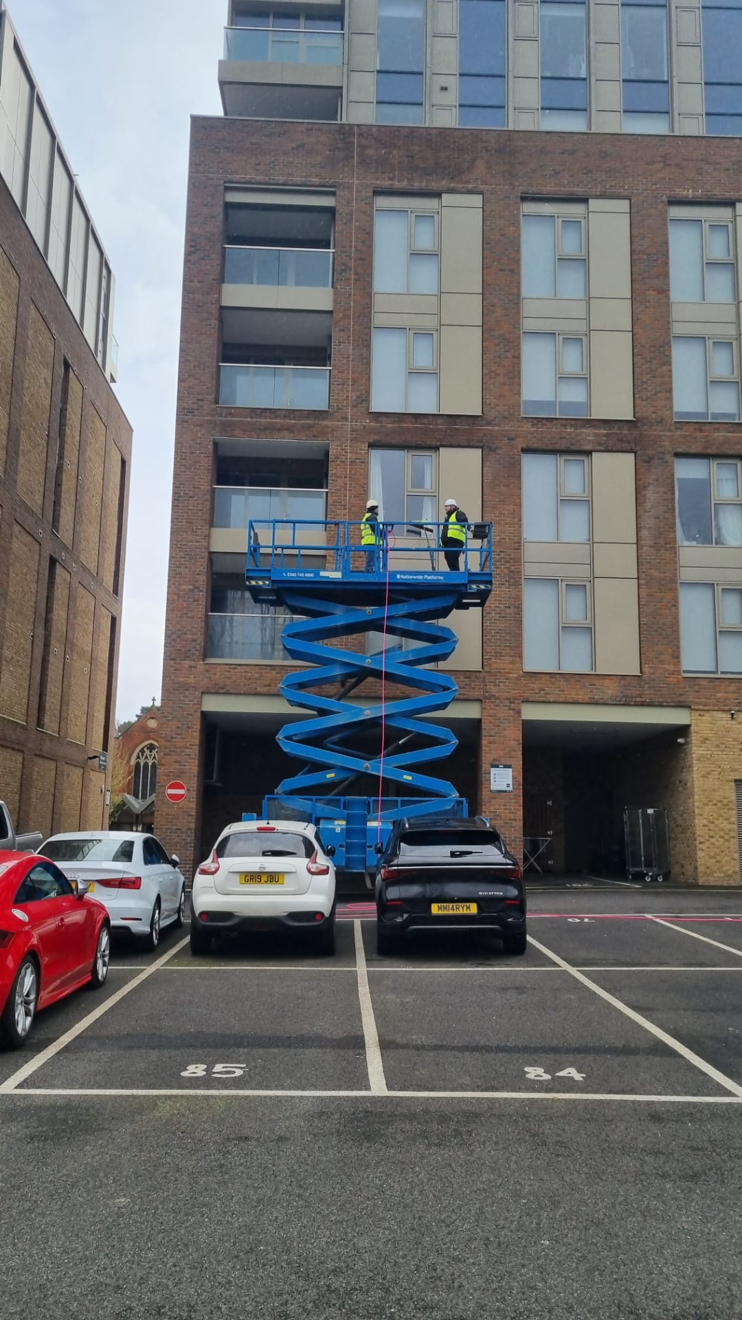 Two operatives in harnesses and helmets cleaning windows from a raised blue scissor lift